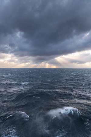 Morning sun rays pierce through dark clouds above the restless sea waves, creating a breathtaking...