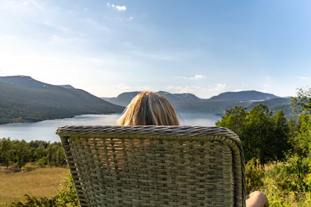 A woman sits on a chair in a garden, watching Gjevillvatnet lake surrounded by mountains in Oppdal Norway. She enjoys her free time in the summer sun and fresh air.の写真素材