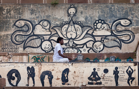 The person practicing yoga in white clothes, sits in a meditation pose near a shiva-lingam, in front of the wall covered with drawings of graffiti. Below, under him, are represented some asanas from yoga complexes. February 6, 2014. Varanasi. India.のeditorial素材