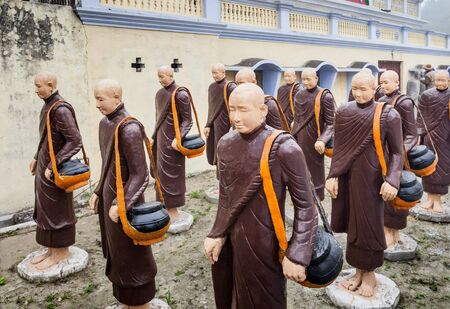 Statues of Buddhist monks, made of concrete, in traditional clothes, with pots for alms, stand at the Thai monastery in the city of Kushinagar.のeditorial素材