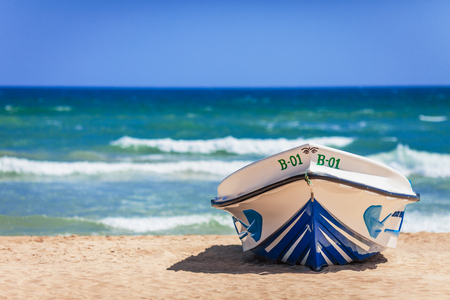 The bright boat of white color with blue inserts, number on the board and with the image of eyes on a nose, is on the sandy coast, against the background of clear sky and blue waves of the southern sea, as reminiscence of hot and bright summer holiday.の写真素材