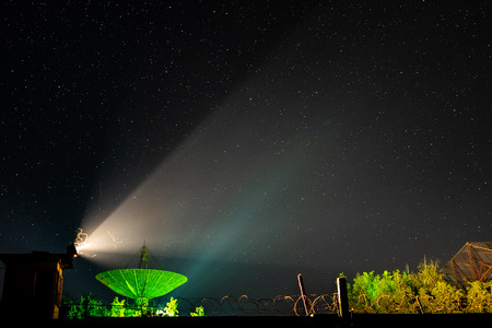 Beam of an old searchlight and line of barbed wire against the background of the big antenna of the radio telescope illuminated by green light under the night star sky.の写真素材