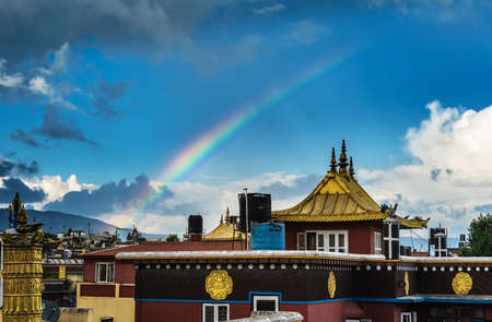Bright rainbow arc over a Buddhist monastery with gilded roofs against the blue sky, dramatic gray clouds and mountains in the background.の写真素材