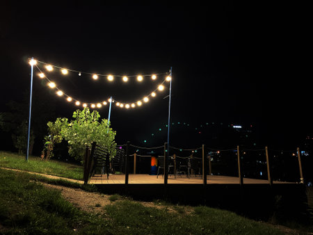 Night terrace, garland lamps, tables on hill overlooking city lights. Wooden platform on shore of pond in Bratislava. Warm glowing decorations. Calm evening lounge photo for tourist or holiday shotsの写真素材