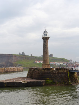 Whitby in England: Stone lighthouse on a harbor pier with a coastal town and green cliffs in the background under a cloudy sky.の写真素材