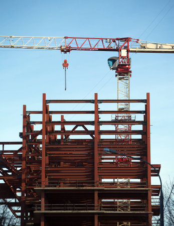 Hoisting tower crane in construction process on top of construction modern building frame over blue...
