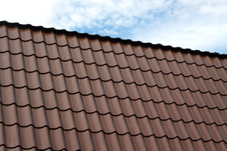 Part of country house roof from brown metal tile under blue sky with white clouds closeupの写真素材