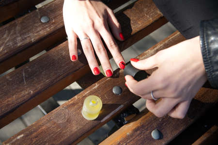 Hands of a young girl who makes a manicure with red nail polish, sitting outside on a bench. Photo...