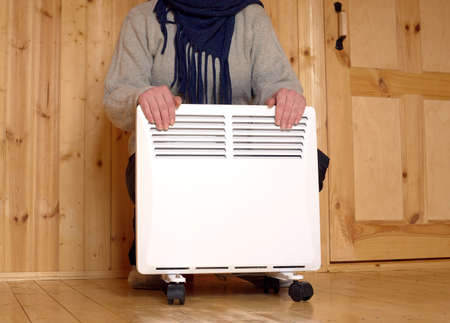 Woman froze and warms her hands on white electric convector heater with horizontal louvers in room of a new wooden houseの写真素材