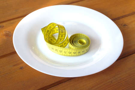 Measuring tape instead food. Measuring tape lies in empty white plate on wooden table surfaceの写真素材