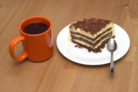 Breakfast. A piece of sponge cake on a white plate with a teaspoon and orange cup of tea on the wooden table surface. Photo closeupの写真素材