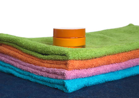 Four clean soft double bath towels set of different colors stacked and jar of cosmetic skin cream on table with denim cloth isoltate on white. Side view from corner close-upの写真素材
