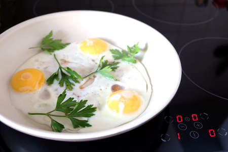 Ready breakfast on stove. Three appetizing fried eggs with parsley in white pan on modern kitchen electric stove with electronic control panel top view closeupの写真素材