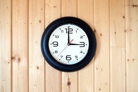 Big round wall clock with a black rim with arrows showing three o'clock hangs on brown wooden wall from vertical planks horizontal view indoor on wooden backgroundの写真素材