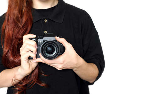 The girl photographer with red hair in a black shirt holds modern black color camera with lens pointing forward. Horizontal indoor studio photo close-up on white background with focus on cameraの写真素材