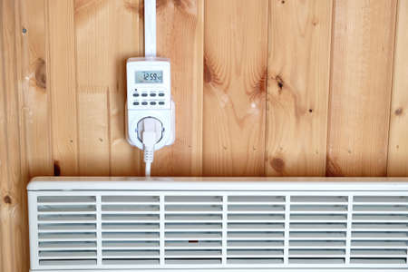 Working white electric convector heater plugged to timer power socket operated in smart house system against wooden wall inside room front view closeupの写真素材