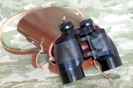 Vintage military Porro prism black color binoculars with brown leather carry case with strap on camouflage background side view close upの写真素材