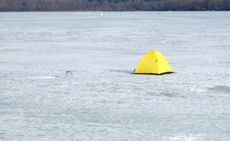 Landscape with fisherman tents in snow on frozen river in winter day against forest at farの写真素材