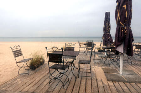 Several wooden tables and chairs on timber platform on empty seaside cafe on Jurmala beach in low season on rainy day view across glassの写真素材