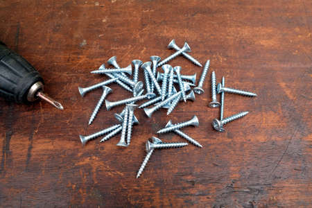 Heap of many light gray screws and old motor screw driver on brown wooden background top view...