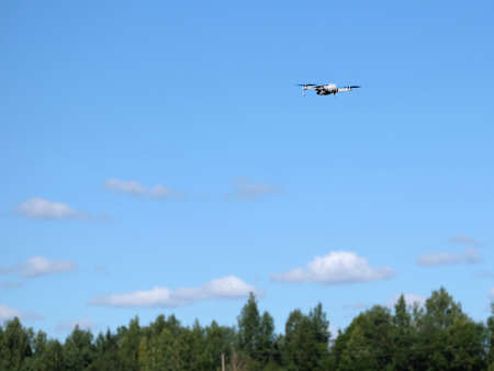 Quadcopter in flight above forest tree tops under blue sky with light white clouds in bright sunny...
