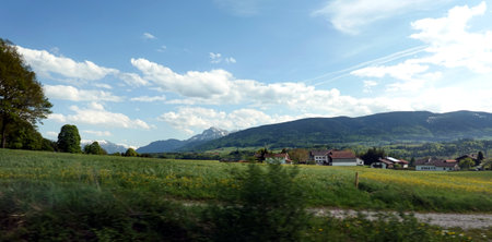 Rural panoramic landscape with typical alpine village on blooming meadows with green grass and high mountains with snow on top at far away, view from fast train window on bright sunny dayの写真素材