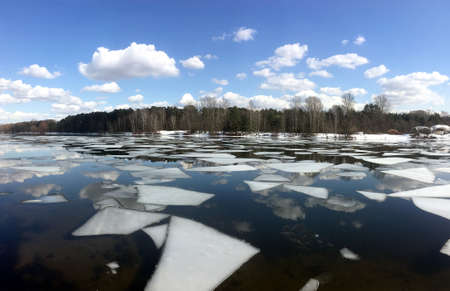 Beautiful natural landscape with ice floes adrift on the spring river and forest trees on opposite bank under clouds on blue sky reflected in the water horizontal viewの写真素材