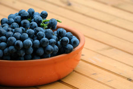 Lot of ripe red grape berries on bunch in brown round bowl on wooden table front view close-upの写真素材