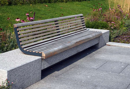 Empty brown wooden bench on stone bases in the city park against green lawn with flowers side view close upの写真素材