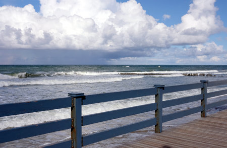 View to the sea waves under blu sky with dense clouds on skyline from embankment with blue wooden fence on sunny windy dayの写真素材