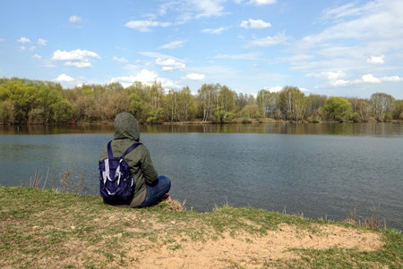 Lonely sitting woman rests on the high lake coast in spring day view from the backの写真素材