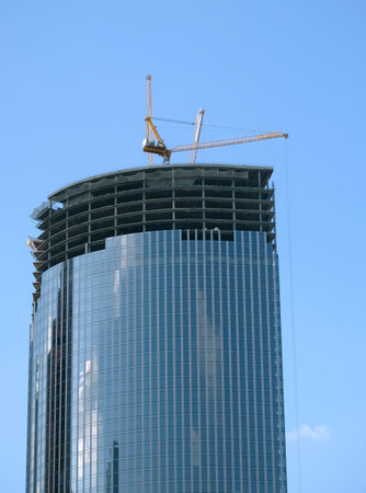 Yellow hoisting tower crane on the top section being construction of modern high skyscraper building against blue cloudless skyの写真素材
