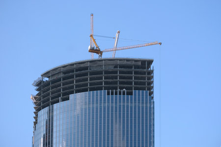 Yellow hoisting tower crane on the top section being construction of modern high skyscraper building against blue cloudless skyの写真素材