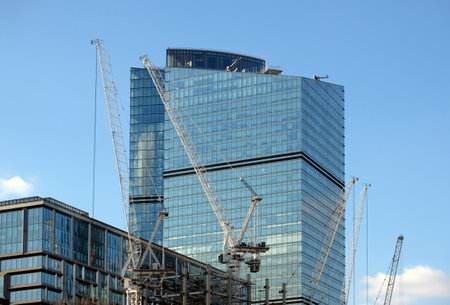 Yellow hoisting tower crane in construction process of constructing skyscraper over blue sky with cloudsの写真素材