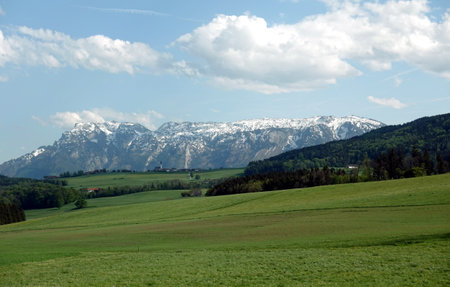 Beautiful rural landscape with blossoming alpine meadows with green grass and mountains with snow caps at far away under blue sky with clouds on bright sunny dayの写真素材