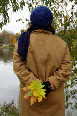 Young girl wear in autumn cotton coat holds behind back bouquet from colorful maple leaves and looks to the river surface from the shoreの写真素材