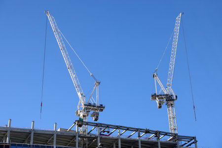 Two hoisting tower crane on the top section being construction of modern high skyscraper building against blue cloudless skyの写真素材