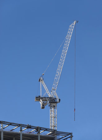Hoisting tower crane on the top section being constructed of modern high skyscraper building against blue cloudless skyの写真素材