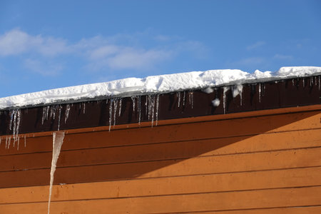 Many melting icicles with fall down drops hangs down close-up on the flat roof with snow against blue spring skyの写真素材