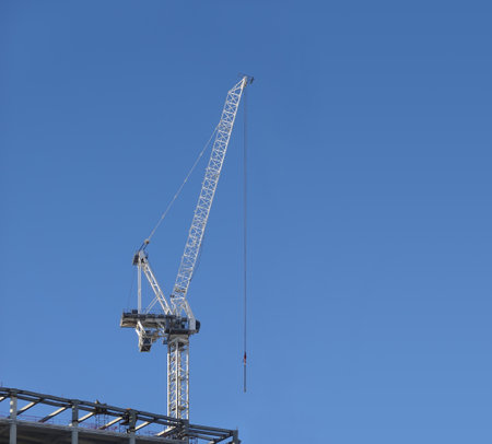 Tall hoisting tower crane on the top section of being constructed of modern high skyscraper building against clear blue cloudless skyの写真素材
