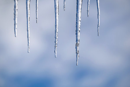 Long crystal icicles hanging down close-up before blurred blue sky with white fluffy clouds in spring dayの写真素材