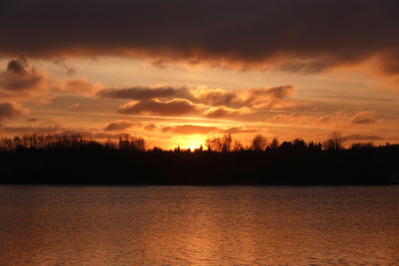 Beautiful natural landscape with dramatic sunset and forest silhouette on horizon against calm riverの写真素材