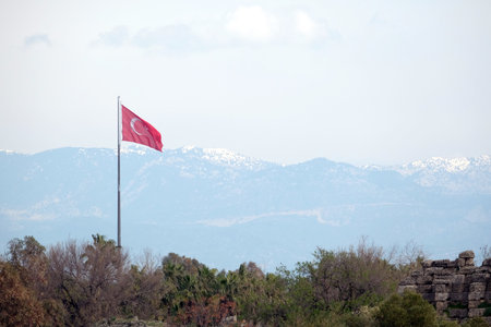 Turkish state flag waving on flagpole before rural landscape with high mountains with snow on the tops at far in light hazeの写真素材