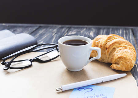 French croissant, glasses, cup of hot coffee, kraft paper and notebook on wooden table. Workplace and Leisure conceptの写真素材