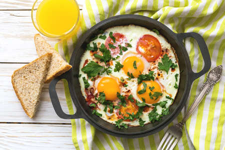 Pan of fried eggs, tomatoes and parsley with bread  and orange juice on white wooden table....