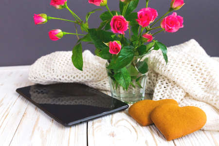 Warm knitted plaid, tablet and heart shape cookies on wooden table. Bouquet of pink roses. Autumn...