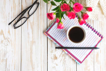 Warm knitted plaid, glasses, cup of coffee, notebook and pensil on wooden table. Bouquet of pink roses. Autumn and winter, leisure concept.の写真素材