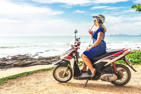 Koh Lanta Island, Krabi Province, Thailand - December 10, 2016: touristic blond Girl with smoothie sitting at Honda motobike on the observation point near exotic beachのeditorial素材