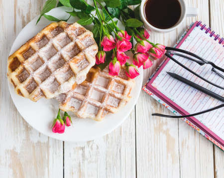 Pink roses, glasses, cup of coffee, notebook, pensil and Belgium waffles on wooden table. Autumn and winter, leisure concept.の写真素材