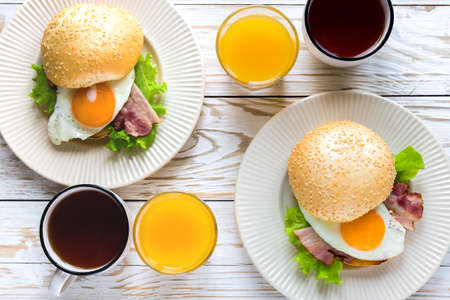 Burger sandwich with sesame bun, orange yolk fried egg, bacon and green salad, cup of tea and orange...
